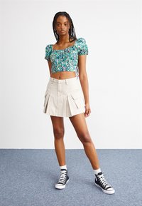 Zöld virágmintás crop top rövid puff ujjakkal, bézs redőzött mini szoknyával, oldalsó zsebekkel és fekete magas szárú tornacipővel párosítva.