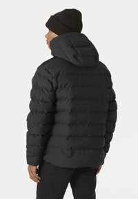 Chaqueta de plumas negra con capucha, que presenta un diseño acolchado y puños elásticos. Llevada con un gorro de punto marrón, que muestra su superficie texturizada.