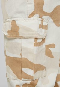 Beige and brown camo cargo shorts featuring a large side pocket with a flap, flat seams, and a smooth cotton fabric texture.