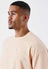 Camiseta de corte oversize en melocotón claro, hecha de algodón suave, con un logo texturizado "PXP" en el pecho y un cuello redondo.