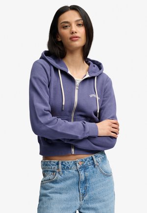 Ung kvinde med glat sort hår iført en lilla hættetrøje med lynlås og lyseblå højtaljede jeans, med armene krydset.