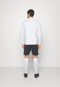 Witte sportshirt met lange mouwen, gecombineerd met donkere shorts, witte sokken en zwarte voetbalschoenen. Eenvoudig ontwerp zonder zichtbare patronen.