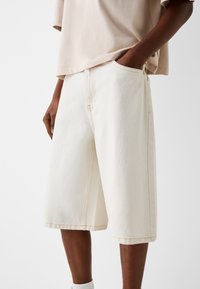 Kermanväriset farkkuhousut, joissa on väljä malli, sivutaskut, näkyvät saumat ja polvimittainen leikkaus. Karheapinta.