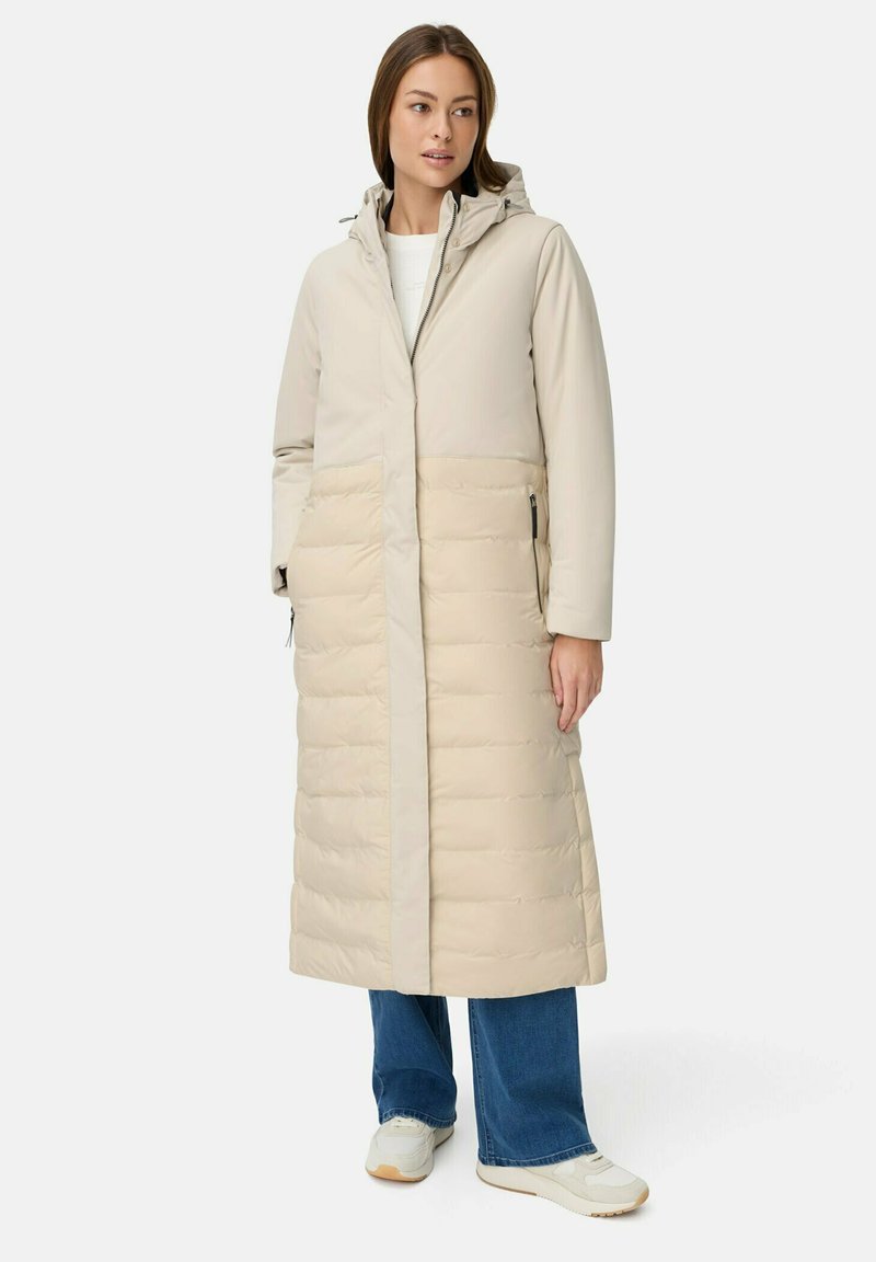 Kvinde iført en lang beige polstret frakke med hætte, blå jeans og hvide sneakers, stående mod en ensfarvet hvid baggrund.