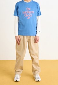 Modré bavlněné tričko s růžovým grafickým nápisem, vrstvené přes bílé tričko s dlouhým rukávem. Kombinováno s béžovými volnými kalhotami a vícebarevnými teniskami.