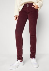 Bordowe spodnie z korduroju o wysokim stanie, z trzema metalowymi guzikami i dopasowanym krojem, w połączeniu z białymi sneakersami.