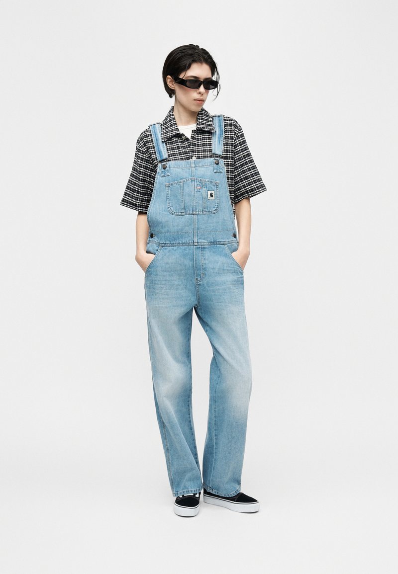 Personne portant une chemise à manches courtes à carreaux noir et blanc sous une salopette en denim bleu clair, chaussures noires sans lacets et lunettes de soleil noires, les mains dans les poches.