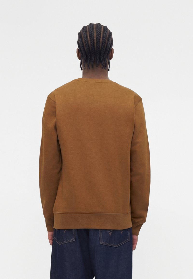 Mann mit geflochtenem Haar, der einen braunen Langarmpullover und dunkelblaue Jeans trägt, steht mit dem Rücken zu einem einfarbigen Hintergrund.