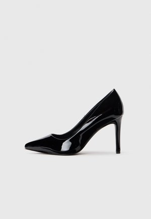 Sort laklæder pumps med høje hæle, spids tåsko og slank stiletthæl mod en lys grå baggrund.