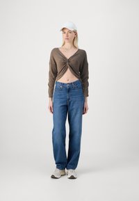 Haut court marron avec un détail torsadé, jean bleu taille haute et baskets blanches. Le mannequin porte une casquette blanche et se tient devant un fond uni.