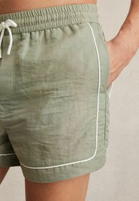 Šviesiai žali maudymosi šortai, pagaminti iš lygaus audinio, su elastingu juosmeniu, raišteliu ir balta apdaila šoninėse kišenėse.