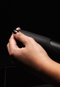 Mano che stringe un dispositivo nero opaco con superficie liscia. La mano ha unghie scure lucidate. Lo sfondo è scuro.