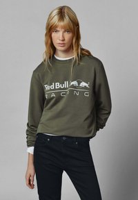 Olive drabužių spalvos megztinis su baltu "Red Bull Racing" logotipu, apvalia kaklo iškirpte ir ilgomis rankovėmis, derinamas su juodais džinsais.