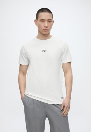 Vyras stovi rankas kišenėse, dėvi baltą marškinėlį su mažu logotipu ir pilkas kelnes, ant paprasto balto fono.