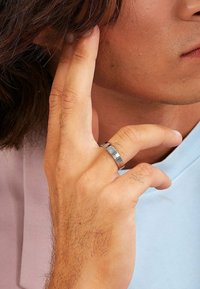 Närbild av en hand med en silverring som gör en gest nära örat, delvis synlig en persons ansikte och axel i en ljusblå skjorta.