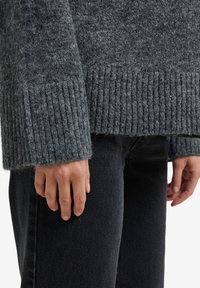 Nahaufnahme von Händen, die entspannt an den Seiten liegen und einen grauen gerippten Pullover sowie schwarze Jeans tragen, vor einem weißen Hintergrund.