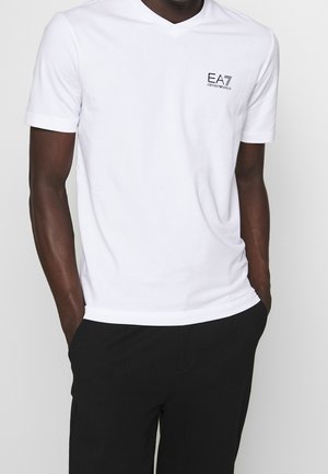 Uomo che indossa una maglietta bianca a maniche corte con scollo a V e logo EA7 Emporio Armani sul petto, pantaloni neri, mani in tasca, su sfondo neutro.