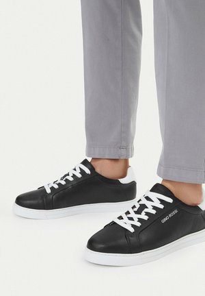 Zapatillas negras de corte bajo con suelas y cordones blancos, usadas con pantalones grises, con la marca "GINO ROSSI" en el costado.