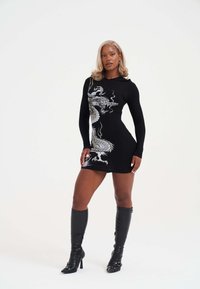 Schwarzes, gestreifrüdes Langarmkleid mit einem weißen Drachendesign. Kombiniert mit knielangen schwarzen Stiletto-Stiefeln mit Schnallen. Glatte Textur.