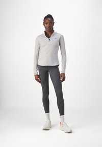 Haut de sport à manches longues gris clair avec un col demi-zip, associé à un legging gris foncé et des baskets blanches avec des accents oranges.