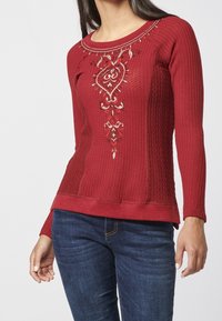 Pull rouge à manches longues avec un motif texturé, présentant des motifs brodés complexes en or et blanc sur le devant.