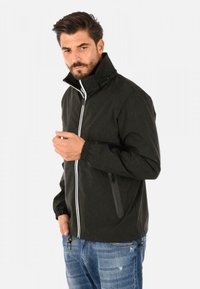 Giubbotto leggero nero con colletto alto e dettagli in zip bianca, dotato di polsini elasticizzati e una tasca laterale, indossato sopra jeans blu.