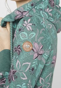 Felpa con cappuccio in tessuto teal, caratterizzata da motivi floreali in lavanda e viola scuro. Include una toppa di marca marrone sulla manica.