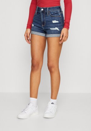 Persoon draagt donkerblauwe verweerde denim shorts met omgevouwen zomen, een rood top met lange mouwen, witte sokken en witte Nike sneakers.