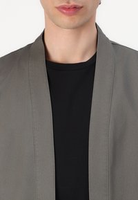 Grå lett jakke med en jevn tekstur, åpen front og uten synlige lukninger, båret over en svart t-skjorte med rund hals.