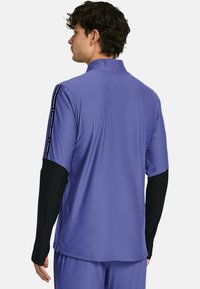 Lila sportliches Kurzarmshirt mit hohem Kragen, schwarzen langärmligen Einsätzen und Under Armour Logo-Akzenten an den Schulternähten.