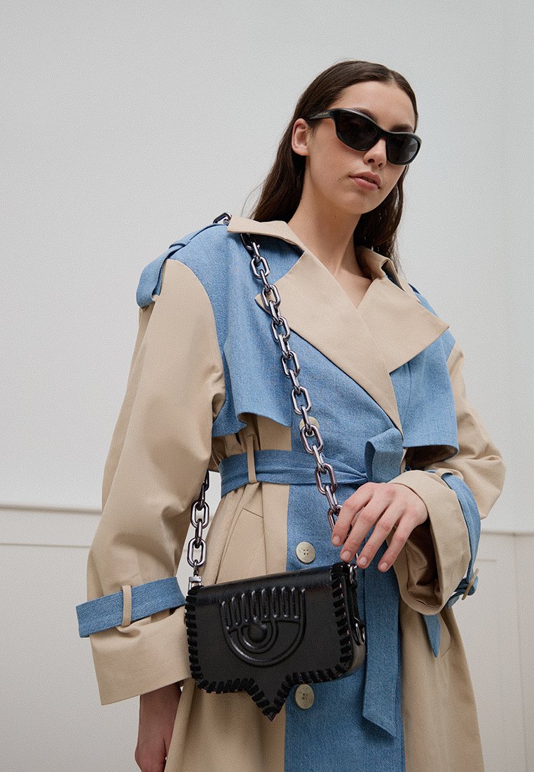 Trench-coat bicolore beige et bleu avec un large col, une taille cintrée, et un sac à main noir avec bandoulière en chaîne arborant un motif décoratif d'yeux.