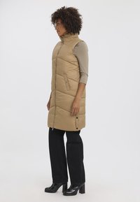 Gilet long rembourré beige avec col montant, poches latérales et motif matelassé, porté sur un haut marron clair et un pantalon noir avec des talons.