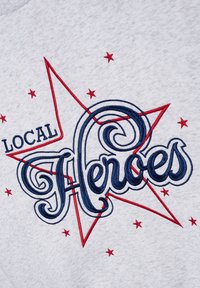 Szara tkanina z czerwonym i niebieskim haftowanym wzorem, na którym znajduje się słowo "LOCAL" nad kursywnie zapisanym "Heroes", umieszczonym w kontekście motywu promieni słonecznych z małymi czerwonymi gwiazdkami.