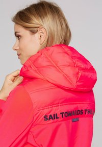 Bright pink, padded jacket with a detachable hood. Features black text on the back reading "SAIL TOWARDS THE" and logo accent.