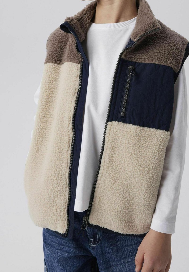 Enfant portant un gilet en polaire beige et marron avec une poche poitrine matelassée bleu marine sur une chemise blanche à manches longues et un jean bleu.