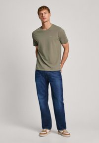 T-shirt verde oliva a maniche corte con collo rotondo, abbinato a jeans blu e sneakers beige. Dettaglio del logo minimal sul petto.