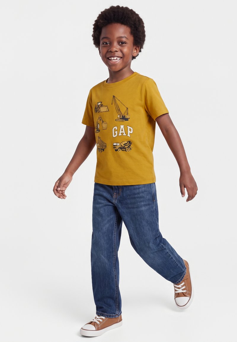 Enfant souriant aux cheveux bouclés portant un t-shirt jaune moutarde avec des engins de chantier et un jean, marchant avec un pied en avant.