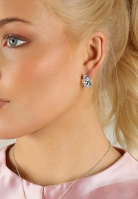 Silverfärgad hängörhänge med vriden design och blommiga detaljer, bärs i vänster öra. Slät yta mot en ljus hudton.