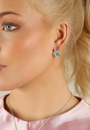 Silverfärgad hängörhänge med vriden design och blommiga detaljer, bärs i vänster öra. Slät yta mot en ljus hudton.