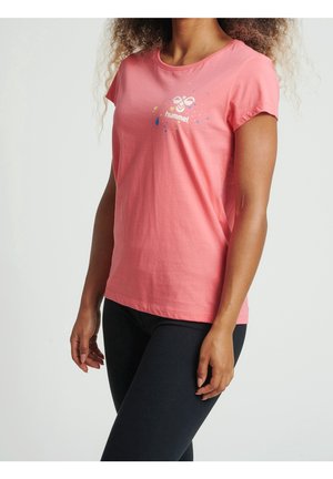 Kobieta ubrana w koralowo-różowy t-shirt z krótkim rękawem z małym białym logo Hummel i plamami koloru, połączona z czarnymi legginsami.