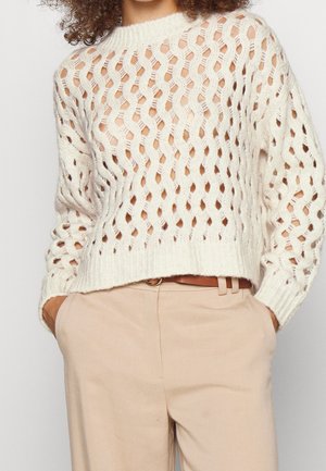 Witte opengebreide trui gedragen over de huid, gecombineerd met beige broek en bruine riem, handen in de zakken gestoken, bij de borst afgesneden.