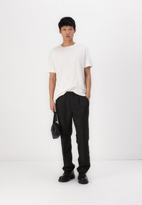 T-shirt en coton blanc, pantalon noir fuselé, chaussures noires épaisses et un petit sac bandoulière noir. Le mannequin se tient devant un fond blanc.