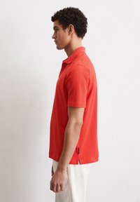 Rode polo shirt met korte mouwen en kraag, een gladde textuur, zij-splitten aan de zoom en een subtiele merketiket aan de onderzijde.