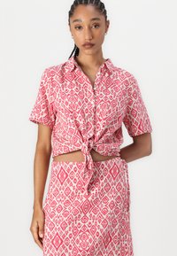 Chemise à manches courtes rouge et blanche à motif, avec un devant noué, dotée d'un col et de motifs géométriques sur l'ensemble.
