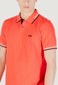 Polo rosso con logo "BOSS" nero, dotato di colletto classico e accenti a righe sulle maniche. Realizzato in tessuto liscio.