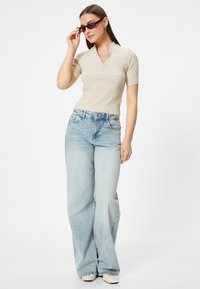 Maglione a coste beige chiaro con colletto zip, abbinato a pantaloni a gamba larga blu chiaro a vita alta. Modello con occhiali da sole scuri.