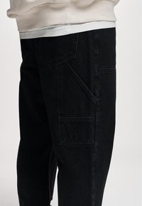 Black denim pants featuring a straight-leg fit, multiple pockets, and a textured surface, with minimal stitching and a clean design.