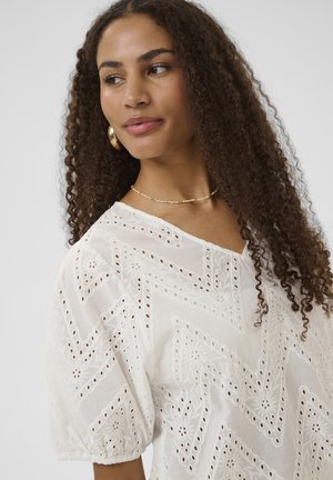 Fiatal nő göndör hajjal, arany karika fülbevalókat, finom nyakláncot visel, valamint fehér, lyukacsos mintájú blúzt fodros ujjakkal.