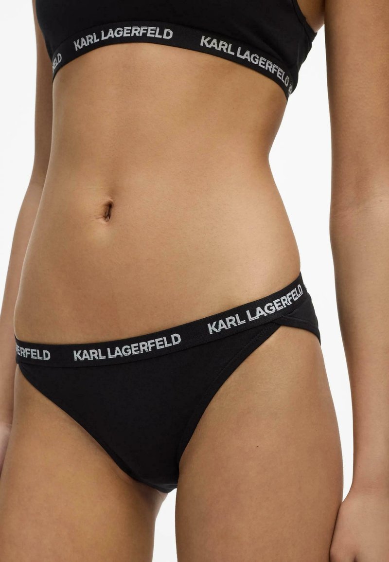 Gros plan d'une personne portant un ensemble de sous-vêtements noirs de la marque Karl Lagerfeld, montrant le logo sur les bandes élastiques du soutien-gorge et de la culotte.