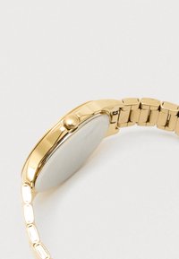 Χρυσής απόχρωσης μέταλλο ρολόι με λεία στρογγυλή κάσα, κρεμ ρουκέτα και συνδεδεμένο λουρί με ορθογώνιες αλυσίδες.